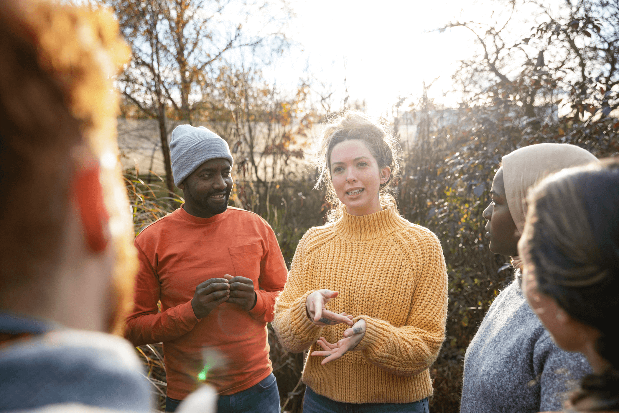 Young woman wearing the yellow jumper is giving directions to a group of volunteers outside in daylight