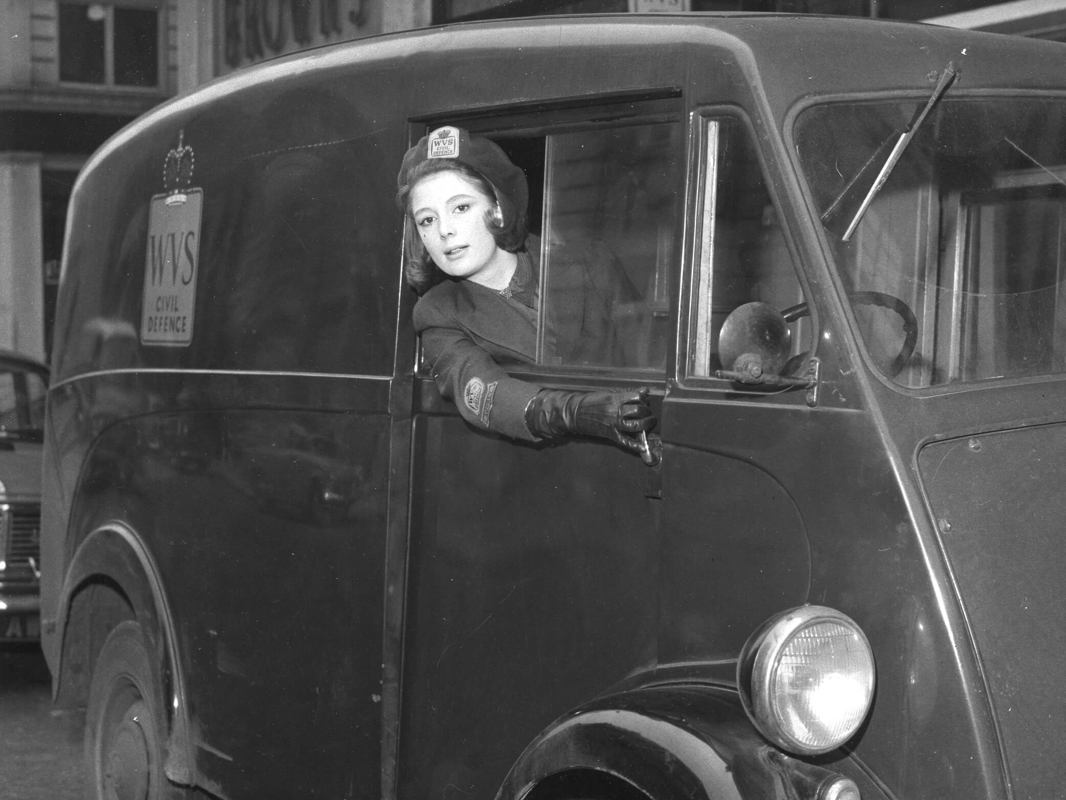 An black and white image of a woman wearing WVS uniform and looking out the drivers side window of a vehicle during the 1940s