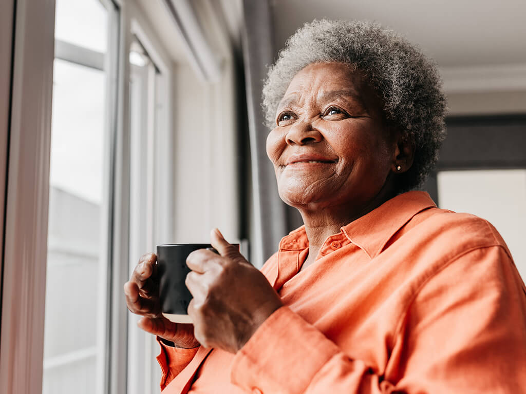 An older woman smiling while looking out of the window and enjoying a hot drink. 