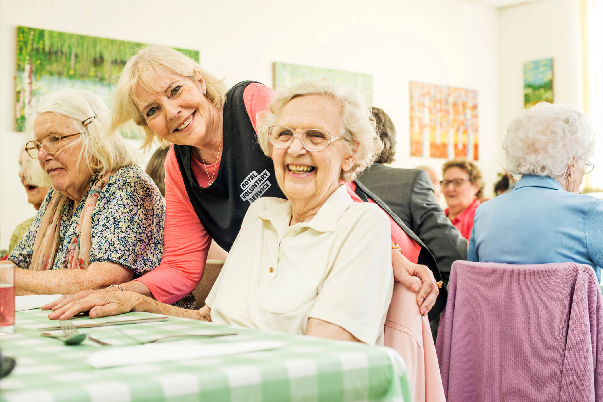 A Royal Voluntary Service lunch club volunteer smiling at the camera with an older person who is sat at a dinner table 